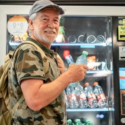 Smiling adult senior man with cap and backpack choosing drinks on 24h free automated distributor - people buying food on airport gate - travel lifestyle